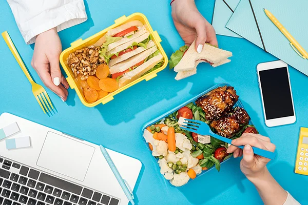 cropped view of two women holding forks over lunch boxes with food near laptop